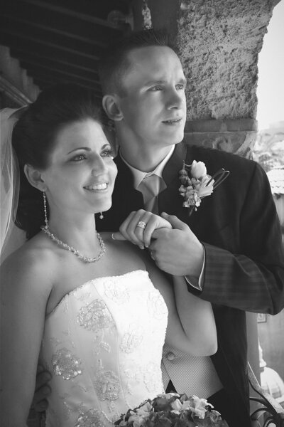 Groom embraces his bride on a balcony on their wedding day as they look off into the distance