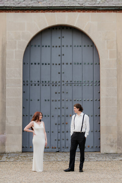 beautiful couple during their engagement session. Women in white dress looking at man in front of large  blue door.