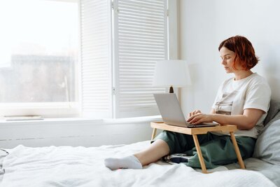 A woman sits in bed, leaning her back against her pillow. She has a small lap desk above her legs, one of which is a prosthesis. The desk supports a laptop, which is types on.