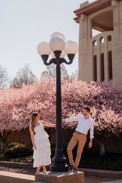 A couple poses under a blooming tree holding a light pole and smiling at each other.