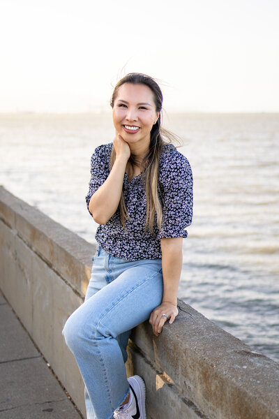 Lifestyle portrait of a Ellen Lai photographer smiling softly as she sits by the water during golden hour, captured by Ellobelle Photography Family Photographer
