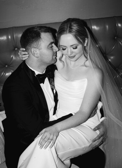 Bride sits on the grooms lap while he nuzzles his nose into her cheek. By Casey McMurray Photography