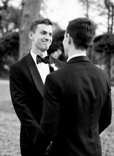 Two grooms in suits stand facing each other outdoors. The person on the left is smiling.