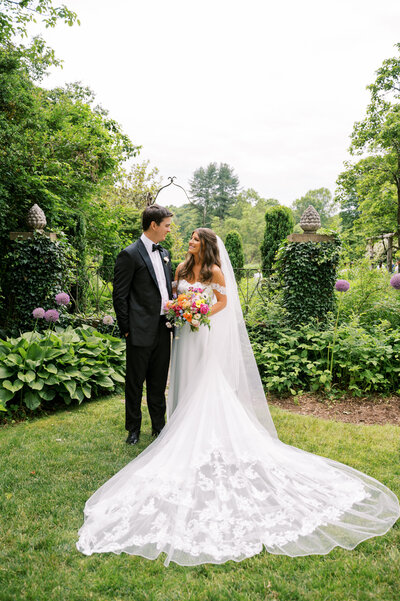 Bride and groom standing in a lush garden, exchanging loving glances, with the bride holding a bouquet and wearing a flowing wedding gown.