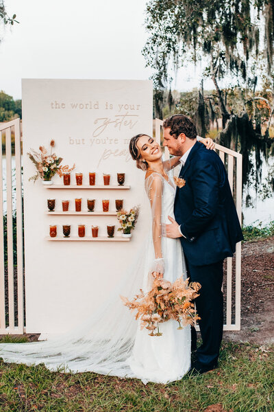 The 'Malibloom' backdrop by Shindig Social, featuring a white panel with the phrase 'the world is your oyster but you're my pearl' and adorned with flower arrangements. A bride and groom stand in front, creating a romantic wedding setting.