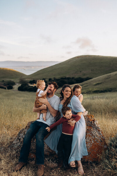 A joyful family of five enjoys a sunset photoshoot outdoors. parents sit on a rock with their three young children, surrounded by grassy hills overlooking a distant body of water.