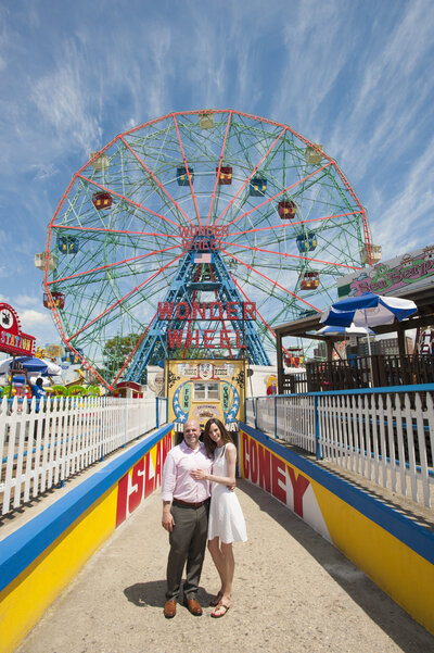 A couple with their arms around each other standing in front of a ferris wheel on Coney Island 