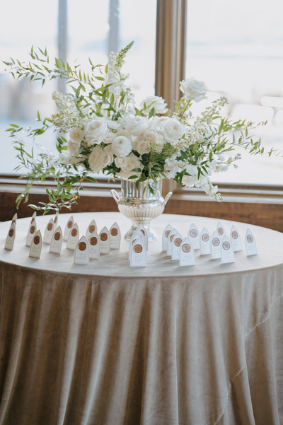An elegantly styled escort card table draped in tan suede linen, featuring a lavish white floral arrangement and Scottish heritage escort cards adorned with gold wax seals, showcasing sophisticated invitation ideas for a refined wedding.
