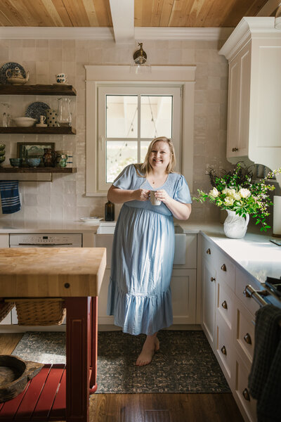 Woman in a light blue dress standing in a kitchen holding a coffee mug
