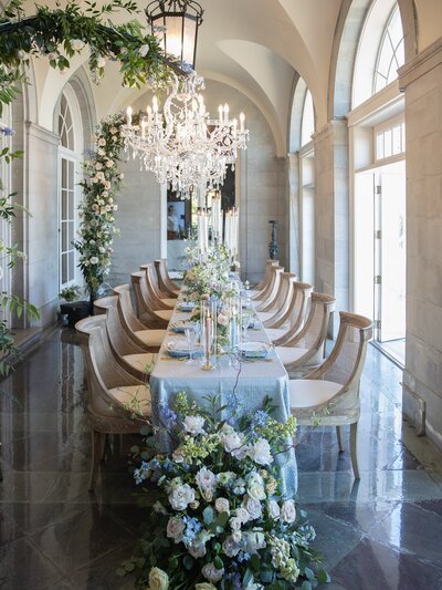 Rhode Island Wedding Photographer | Kelsey Sheehan Photography Timeless Rhode Island Weddings | Elegant dining room with a long table set for guests. Crystal chandelier, floral centerpiece, arched windows, and lush greenery create a sophisticated ambiance.