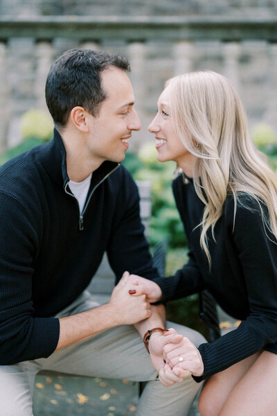 Engaged couple holding hands and smiling at each other during an intimate engagement session outdoors in soft natural light