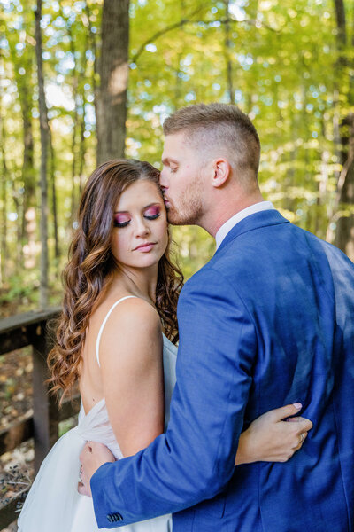A bride looks at the ground while her groom kisses the side of her face. He is in a blue tuxedo and she has on pink eyeshadow.