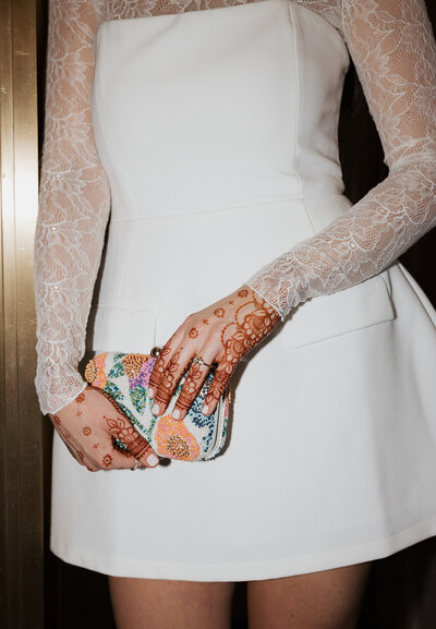 Bride in short white dress with lacy sleeves holds colorful clutch and shows off engagement ring with detailed henna hands.