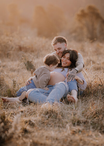 Couple sitting in a field . Dad is kissing newborn while mum rests against dad's back.