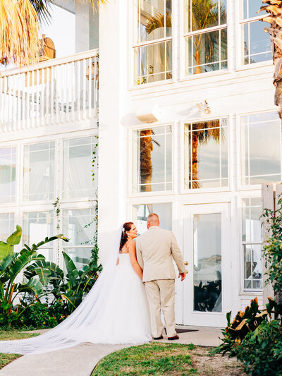 A bride and a groom walk towards their wedding venue. The venue is a tropical venue, featuring an all-white building with plenty of windows and a balcony looking over the water. You can see the reflection of a palm tree in the windows. The bride is wearing a cathedral length veil and a strapless wedding dress, and the groom is wearing a tan suit. The image was taken by a company that specializes in wedding photography in San Antonio.