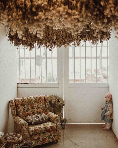 Toddler standing in the corner of a white room with dried flowers covering the ceiling.