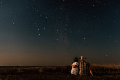 Kate Simpson and her partner sit together under a starry night sky, gazing at the stars in a peaceful, wide-open landscape.