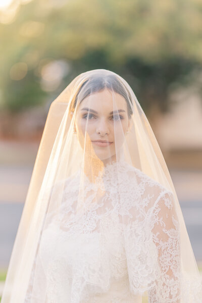 A bride gazes softly through her delicate lace veil, bathed in golden light, capturing a classic and romantic wedding moment
