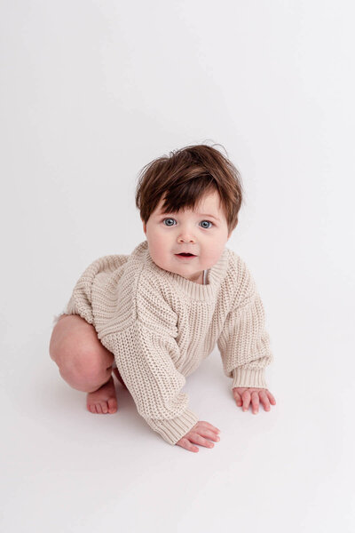six month old baby in cream romper and brown beanie sitting up and leaning towards camera.