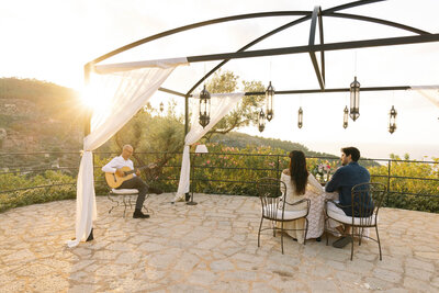 A couple sits at a table under an open canopy adorned with lanterns and white drapes, overlooking a scenic mountain view. A musician plays a guitar nearby. The sun sets in the background, casting a warm glow over the scene.