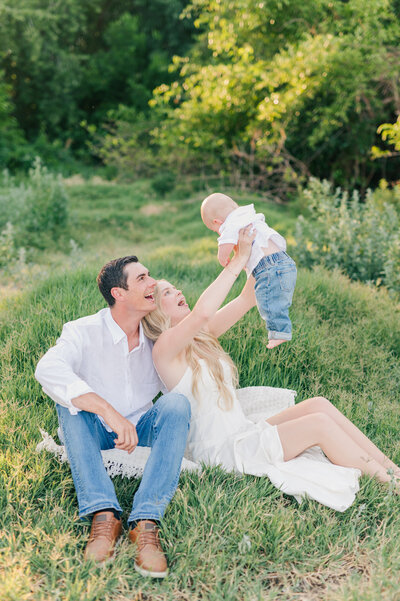 A family of three posed sitting with their baby held in the air at a lush green location in Lewiston ID