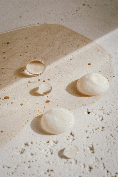 Close-up of a beige surface with spilled liquid and several translucent blobs scattered across it. The surface has small speckles, and the light creates soft shadows around the drops and blobs.