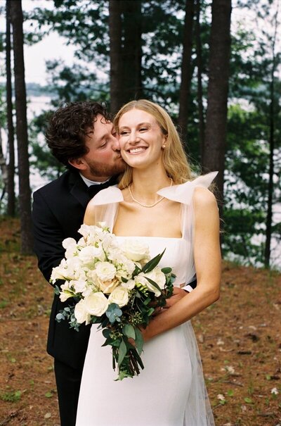 bride and groom embraced outside bride smiles holding bouquet while groom kisses her cheek taken in minnesota by wedding photographer tom thornton
