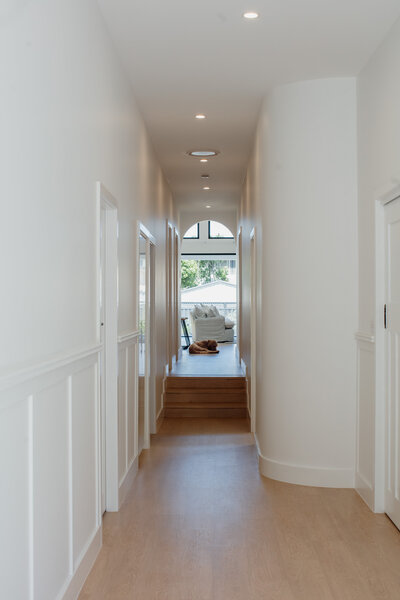 Long, light hallway with wooden floors, embodying the essence of luxury home design in Newcastle, leads to a sunlit seating area with a large window at the end.