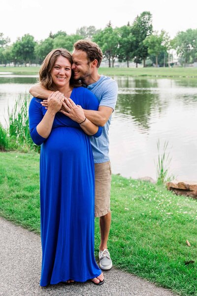 Smiling mom to be in blue dress is embraced by smiling partner