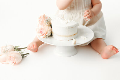 1 year old baby girl sitting with a naked style cake and pink flowers at feet for first birthday cake smash with fresno photographer, one good shot photographer