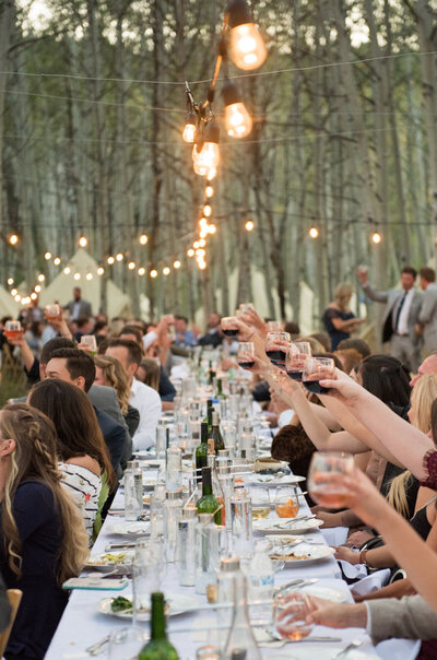 Celebratory wedding toast at an outdoor venue with a family style long table under string lights