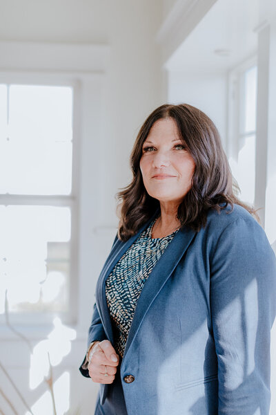 Professional portrait of Susie Schumacher standing confidently in a bright, sunlit room, wearing a navy blue blazer and patterned blouse, with soft natural light illuminating her face - Susie Schumacher Life Coach