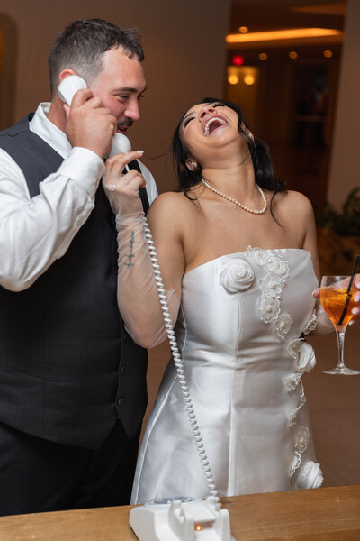 New England Wedding Photographer Pricing | Kelsey Sheehan Photography Timeless Rhode Island Weddings | A bride in a white dress and pearls laughs joyfully while holding a vintage phone with a groom in a suit. Both appear happy, sharing a playful moment.