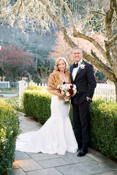 Bride and groom standing on pathway smiling and hugging each other holding a bouquet of red and white flowers