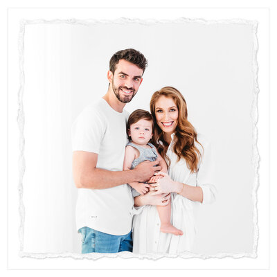 The image depicts a classic family portrait captured in a pristine white studio setting. The focal point is a mother, father, and their young son, all dressed in a harmonious color palette that enhances the serene and timeless atmosphere.  The mother stands elegantly in the center, wearing a flowing white dress that complements her graceful posture. She tenderly holds her toddler son, who is dressed in a light blue jumper, his innocent expression and chubby features radiating a sense of warmth and joy.  Beside them, the father stands tall and proud, his arm gently wrapped around the mother's shoulder. He is dressed in a crisp white shirt and classic blue jeans, his relaxed yet confident stance adding to the overall sense of family unity and stability.  The entire scene is bathed in a pure, classical style, with the stark white backdrop creating a clean and minimalist canvas that allows the subjects to take center stage. The photographer, Bri Sullivan, has masterfully captured the intimate connection between the family members, their expressions and body language conveying a sense of love, comfort, and togetherness.  This family portrait is a testament to the power of simplicity and the enduring beauty of classic portraiture. The clean, pure aesthetic and the harmonious composition create a timeless and elegant image that will undoubtedly be cherished by the family for years to come.