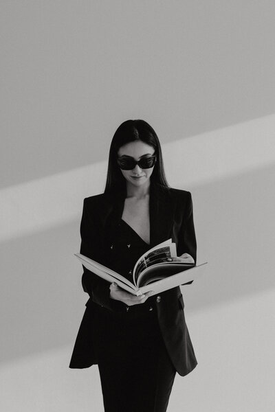 A minimalist black and white portrait of an elegant young woman with black hair, wearing dark sunglasses and holding an open book in front of the camera. Dressed in modern minimalist business attire and haute couture fashion inspired by Dior, she stands against a clean, light background. The image captures cinematic studio lighting with shadows on her face, offering a sophisticated look ideal for luxury branding, custom web design, and high-end branding services. This elegant shot emphasizes creativity, luxury positioning, and modern fashion aesthetics, perfect for bespoke web design and brand identity design services.