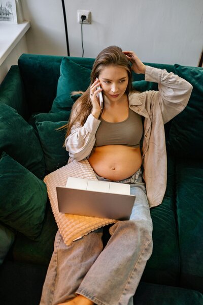 Pregnant woman sitting on couch with laptop and phone during virtual midwifery support
