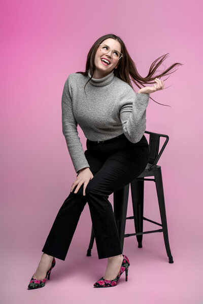 Lana Stewart Photographer sits for self-portrait in studio against pink background, flips hair and smiles over shoulder wearing gray turtleneck
