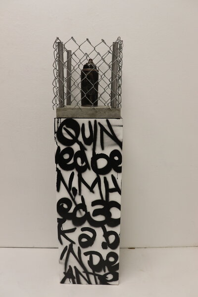 A dark ceramic spray paint can is decorated with a carving of young children and sits atop a graffiti painted pedastal, but is caged in by a wire fence