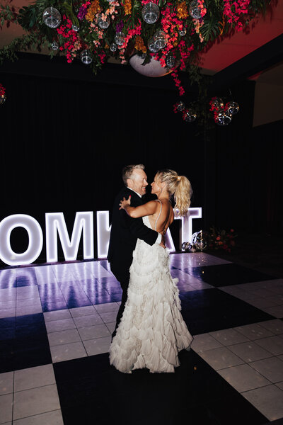 A newlywed couple shares a dance on a black-and-white checkered floor, surrounded by glowing marquee letters and floral decorations with hanging disco balls. Representing Pretty Pop Balloons vendor profile for the Crush Collective wedding show.
