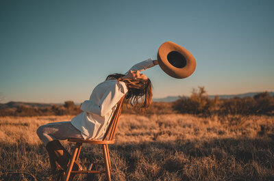 Outdoor photo of Juliet Trnka sitting on a chair with a cowboy hat in one hand. Direct Knowing brand photos