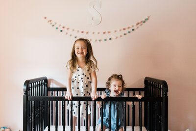 Two joyful children standing in a crib, laughing in a room with pastel-colored walls during their family photos session in Pittsburgh.