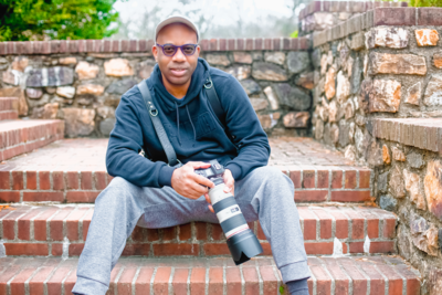 African American man sitting on brick steps with canon long range zoom camera in his hands wearing blue and grey sweats and a baseball cap