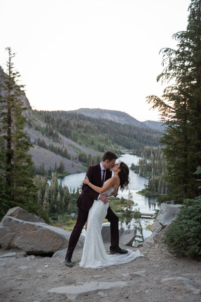 Bride and groom kissing on a rocky overlook during their Mammoth Lakes, California elopement, with alpine trees and a mountain lake in the background.