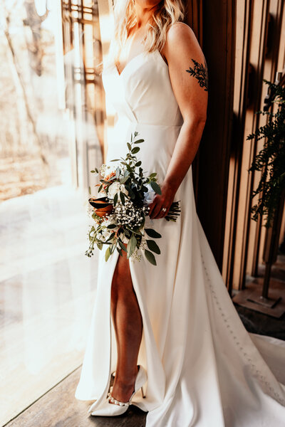 Details of the bride's dress, flowers, and shoes as she stands by the large  glass window of the glass chapel in Northwest Arkansas