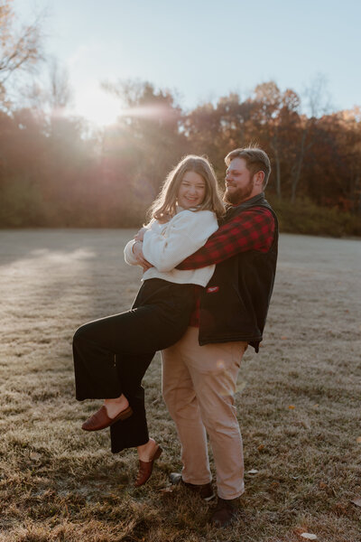 A couple joyfully embraces in a sunlit park. The man, wearing a red flannel, lifts the woman, who smiles in a white sweater. Autumn trees and warm sunlight provide a serene background.