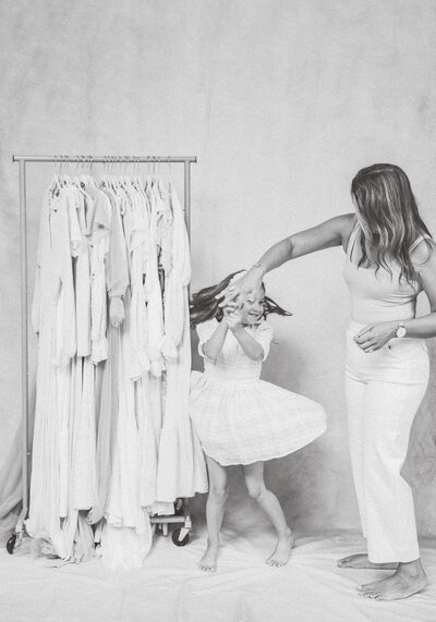 other and young daughter dancing joyfully beside a rack of neutral-toned dresses, captured in a black-and-white studio portrait.
