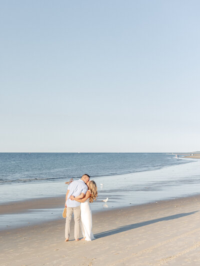 Couple holding hands and kissing while standing barefoot in the sand in front of the ocean