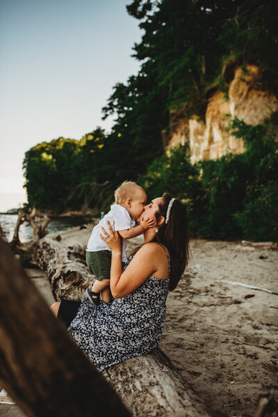 Family photographers Maryland captures mommy and me session with mother holding and kissing her toddler as they sit on a log on the beach