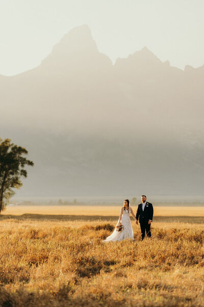 bride and groom walking through an open field with mountains in the background